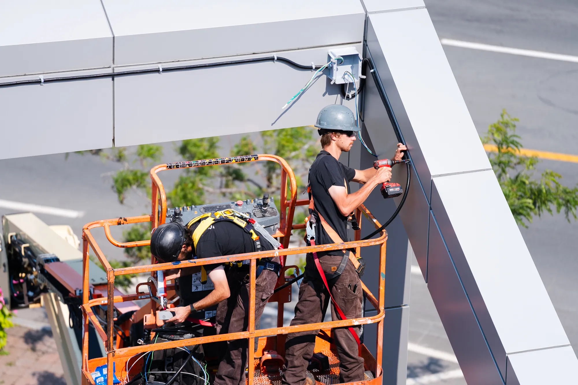 Two LuMar Electric electricians working from an aerial lift basket, running conduit into a junction box beneath a building canopy